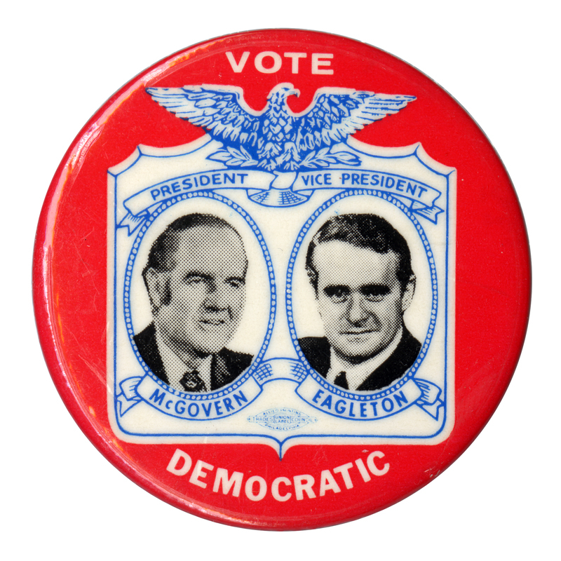 Round, red button with black and white portraits of George McGovern and Sargent Shriver in an elaborate blue and white frame topped by an eagle. Text reads "VOTE DEMOCRATIC PRESIDENT McGOVERN VICE PRESIDENT EAGLETON"