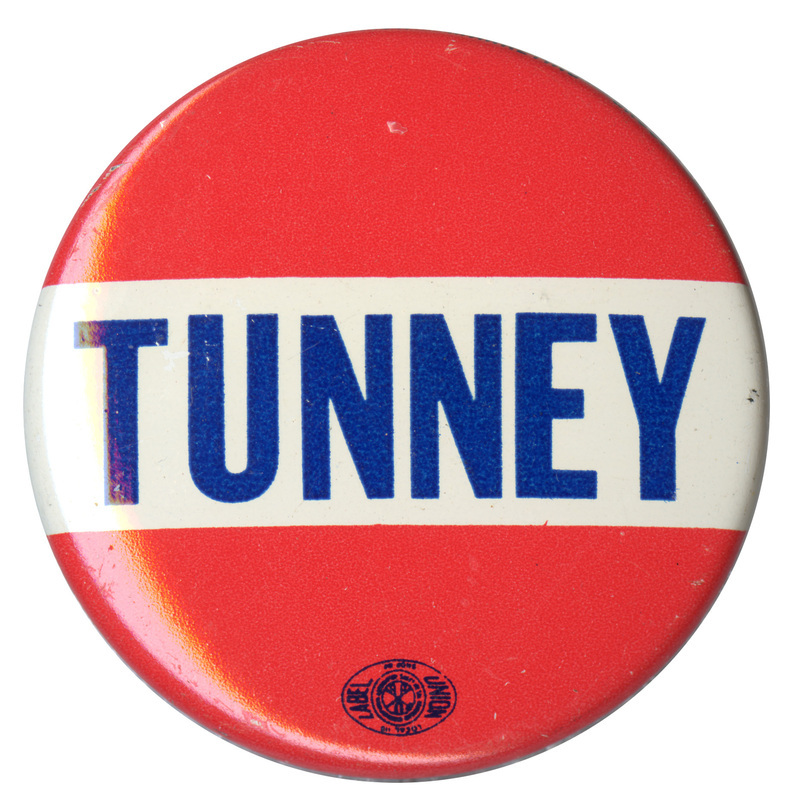 Round, red button with a white band across the middle containing blue text that reads "TUNNEY"
