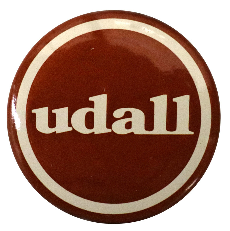 Round, maroon and white button with white text reading "udall"