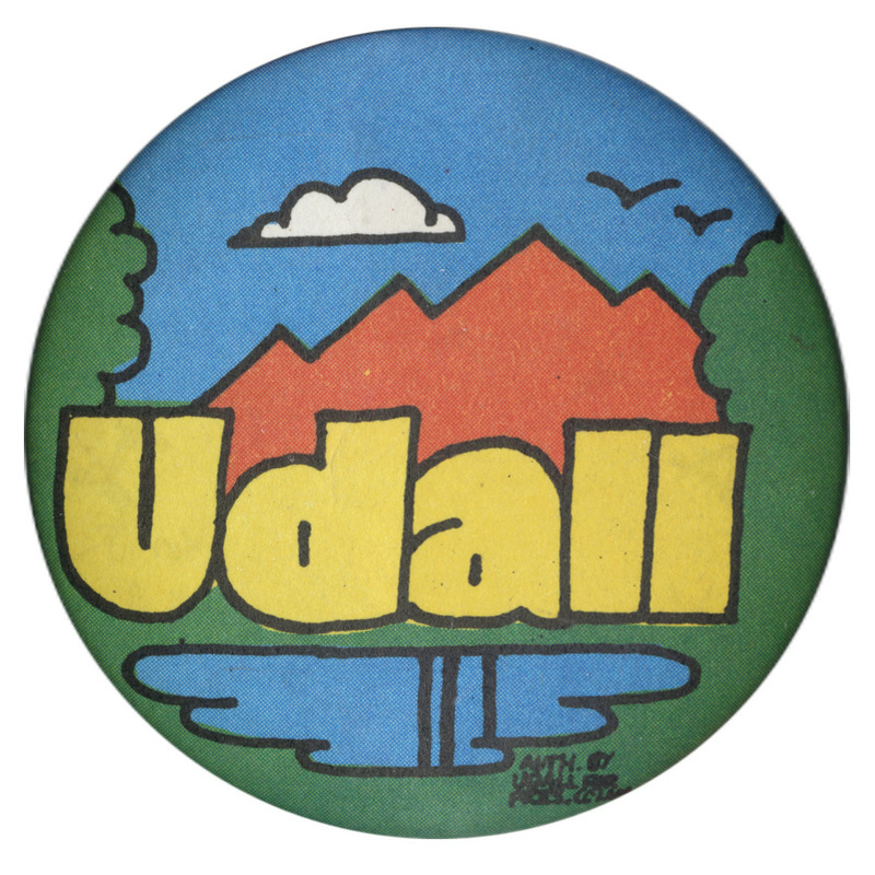 Round button with a green, red, blue, and white drawing of a mountain, trees, and lake. Yellow text reads "udall"
