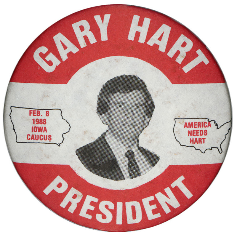 Round, white button with a red border and a black and white photograph of Gary Hart in the center. An outline of the state of Iowa contains red text reading "FEB. 8 1988 IOWA CAUCUS." An outline of the United States contains red text reading "AMERICA NEEDS HART." White text around the edge reads "GARY HART PRESIDENT"