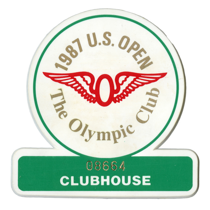 Button in the shape of a circle sitting atop a broad rectangle. Within the white circle is the red, winged symbol of the Olympic Club in San Francisco and gold text reading "1987 U.S. OPEN The Olympic Club." Within the green rectangle is gold and white text reading "08664 CLUBHOUSE"
