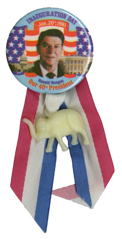 Round button with a color photograph of Ronald Reagan against color photographs of the US Capitol and the White House and US flag decorations. Red, white, and blue text on the button reads "INAUGURATION DAY Jan. 20th, 1981 Ronald Reagan Our 40th President". Attached to the bottom of the button is a red, white, and blue ribbon and a white plastic elephant.