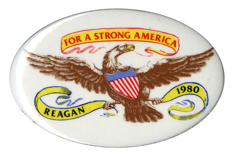 Oval, white button with a color drawing of an eagle bearing a red and blue shield and holding yellow ribbons. Red and black text reads "FOR A STRONG AMERICA REAGAN 1980"
