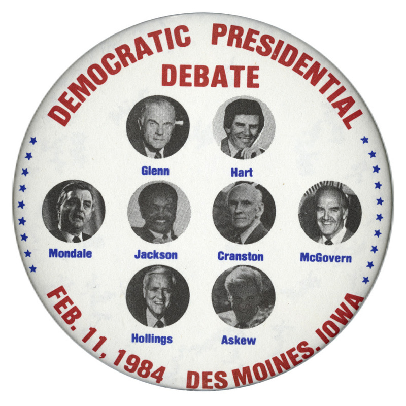 Round, white button with black and white photographs of John Glenn, Gary Hart, Walter Mondale, Jesse Jackson, Alan Cranston, George McGovern, Fritz Hollings, and Reubin Askew, each with his last name underneath his photograph. Red text reads "DEMOCRATIC PRESIDENTIAL DEBATE FEB. 11, 1984 DES MOINES, IOWA"