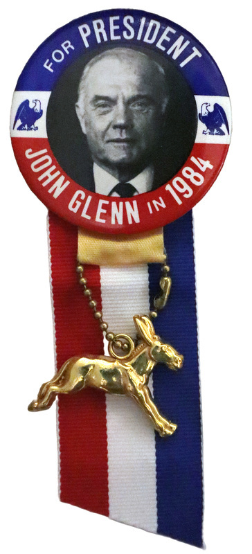 Round button with red and blue border and a black and white photograph of John Glenn flanked by blue eagles. White text reads "FOR PRESIDENT JOHN GLENN IN 1984." Attached to the bottom of the button is a red, white, and blue ribbon and a gold donkey charm.