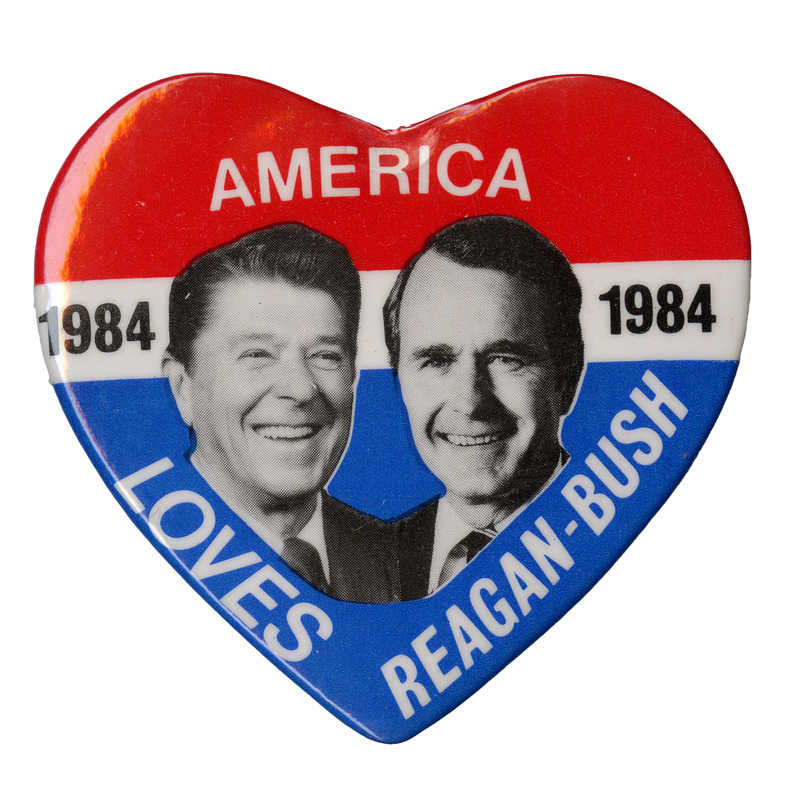 Heart-shaped button with bands of red, white, and blue and black and white photographs of Ronald Reagan and George H. W. Bush. White and black text reads "AMERICA 1984 LOVES REAGAN-BUSH"