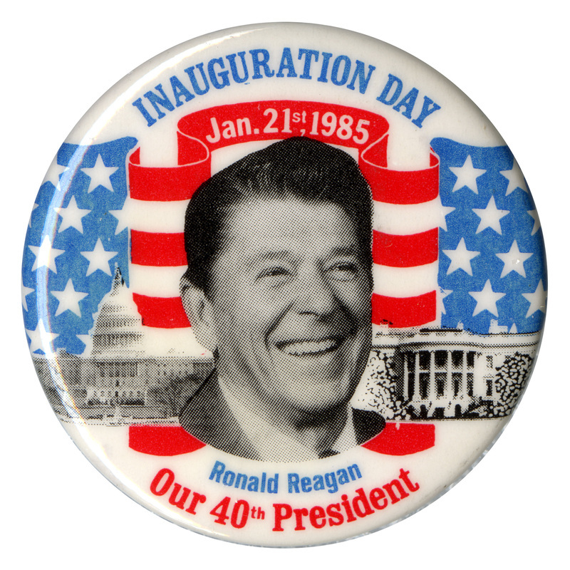 Round, white button with a black and white photograph of Ronald Reagan against black and white photographs of the US Capitol, the White House, and a drawing of the US flag in red, white, and blue. Blue, white, and red text reads "INAUGURATION DAY Jan. 21st, 1985, Ronald Reagan, Our 40th President"