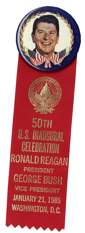 Round button with a blue border with gold leaves and crossed flags around a color photograph of Ronald Reagan. Attached to the bottom of the button is a red ribbon with a gold seal and gold text reading "50th U.S. INAUGURAL CELEBRATION RONALD REAGAN PRESIDENT GEORGE BUSH VICE PRESIDENT JANUARY 21, 1985 WASHINGTON ,D. C."