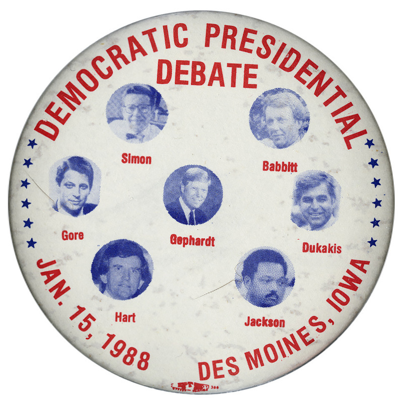 White button with black and white photographs of Paul Simon, Bruce F. Babbitt, Al Gore, Richard A. Gephardt, Michael Dukakis, Gary Hart, and Jesse Jackson, each with his name underneath his photograph. Red text reads "DEMOCRATIC PRESIDENTIAL DEBATE JAN. 15, 1988 DES MOINES, IOWA"