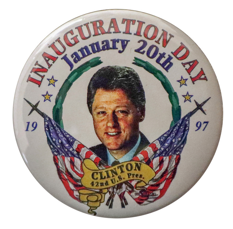 Round, white button with a color photograph of Bill Clinton surrounded by color illustrations of two US flags, greenery, and yellow stars. Red and black text reads "INAUGURATION DAY January 20th 1997 CLINTON 42nd U.S. Pres."