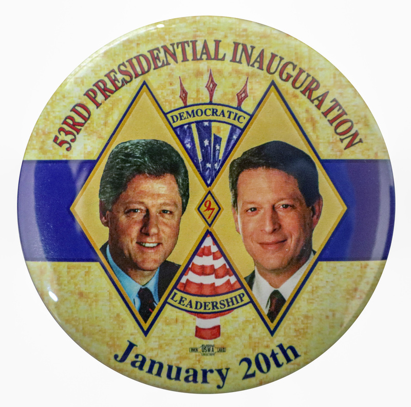 Round button with a splotchy yellow background and a band of blue across the middle. Color photographs of Bill Clinton and Al Gore appear inside interlocked diamond shapes in the center. Between the photographs is an illustration of a US flag. Red and black text reads "53RD PRESIDENTIAL INAUGURATION DEMOCRATIC LEADERSHIP January 20th"