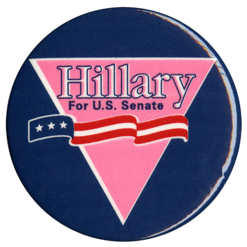 Round, blue button with an inverted pink triangle and a ribbon with red, white, and blue stars and stripes. White and blue text reads "Hillary For U.S. Senate"