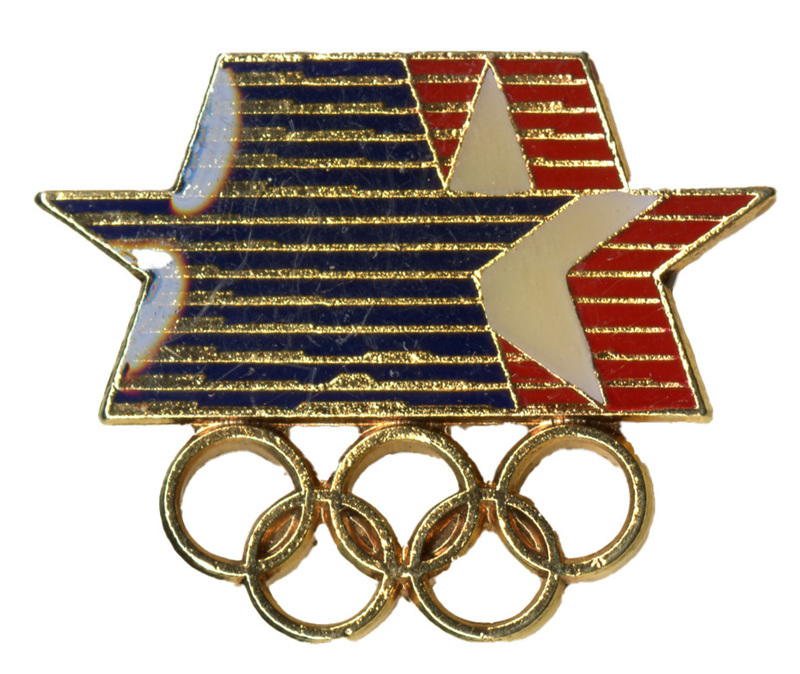 Enameled pin in the shape of linked blue, white, and red stars above five interlocking gold rings. 