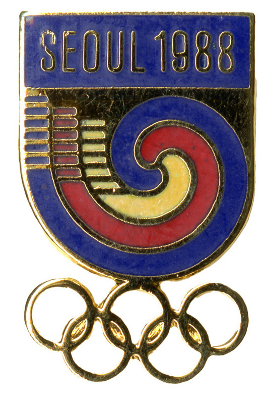 Enameled U-shaped pin with a blue, red, and yellow swirl design below gold text reading "SEOUL 1988". Attached to the bottom of the pin are five interlocking gold rings.