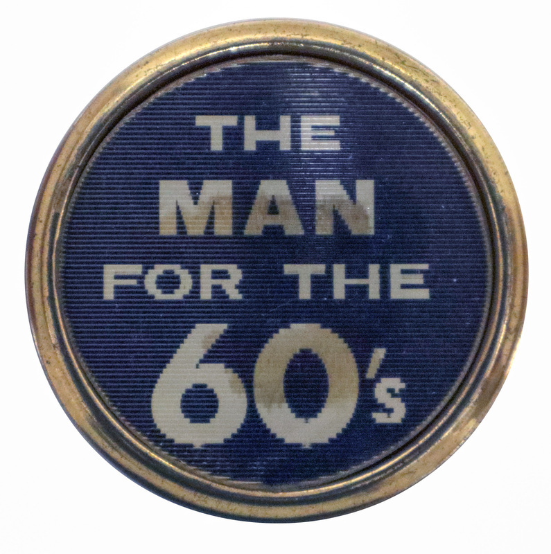 Round button with a gold rim. White text on a blue background reads "THE MAN FOR THE 60'S". This is one angle of a holographic button.