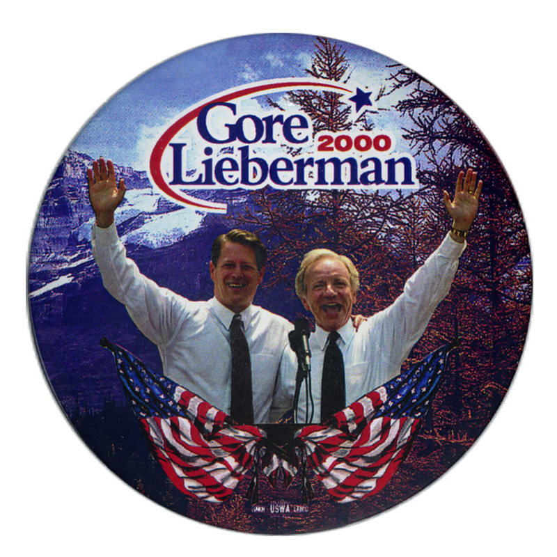 Round button with a color photograph of Al Gore and Joe Lieberman at a podium decorated with illustrations of a US flag. The background is a color photograph of trees and a snow-covered background. Blue and red text reads "Gore Lieberman 2000"