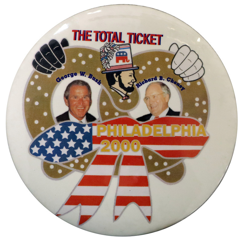 Round, white button with an illustration of one black hand and one white hand grasping a salt-covered pretzel that has a red, white, and blue ribbon attached to it. Within the spaces of the pretzel are color photographs of George W. Bush and Dick Cheney. Above the photographs is a color illustration of a bearded man wearing a tall hat with the red and blue GOP symbol of an elephant. Red, blue, and yellow text reads "THE TOTAL TICKET George W. Bush Richard B. Cheney PHILADELPHIA 2000"