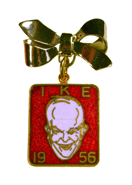 Rectangular button with a gold bow attached to the top. Button features a caricature of Dwight D. Eisenhower on red background. The words "IKE" and "1956" are displayed in gold.