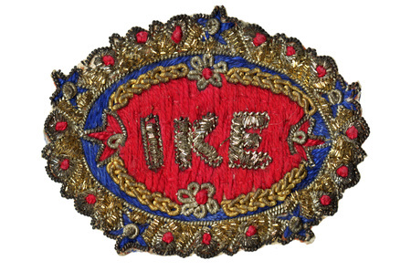 Oval-shaped fabric patch with "Ike" stitched in metallic thread on a red background. Blue and gold floral embroidery border with red accents.