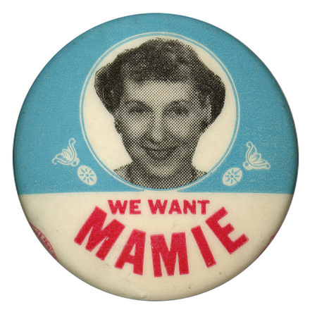 Round, light blue button with a white horizontal stripe at the bottom and a grayscale portrait of Mamie Eisenhower. In the white stripe is red text that reads "We want Mamie". 