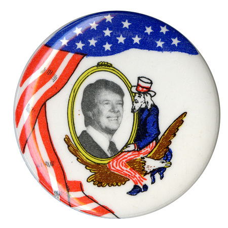Round, white button with a red, white, and blue drawing of the US flag and a drawing of Uncle Sam sitting on an eagle and cradling an oval that contains a black and white photograph of Jimmy Carter.