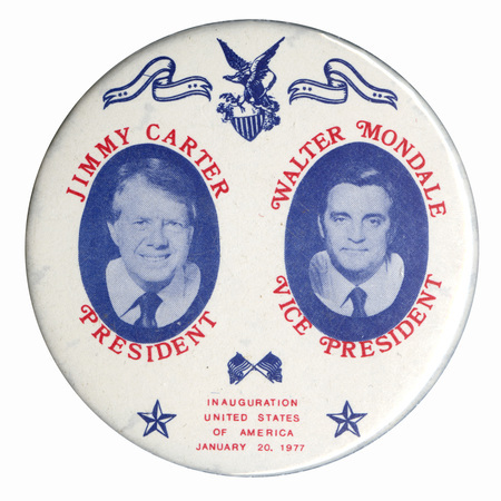 Round, white button with black and white portraits of Jimmy Carter and Walter Mondale, as well as blue drawings of an eagle holding a shield, ribbons, flags, and stars. Red text reads "JIMMY CARTER PRESIDENT WALTER MONDALE VICE PRESIDENT INAUGURATION UNITED STATES OF AMERICA JANUARY 20, 1977"