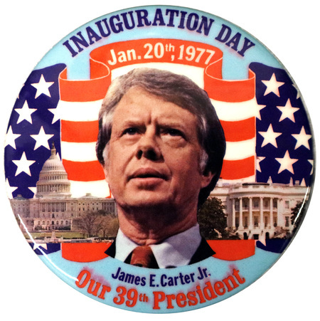 Round button with a color photograph of Jimmy Carter against color photographs of the US Capitol building, the White House, and a US flag. Red, white, and blue text reads "INAUGURATION DAY Jan. 20th, 1977 James E. Carter Jr. Our 39th President"