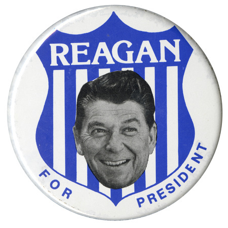 Round, white button with a black and white photograph of Ronald Reagan's head against a drawing of a blue shield. White and blue text reads "REAGAN FOR PRESIDENT"