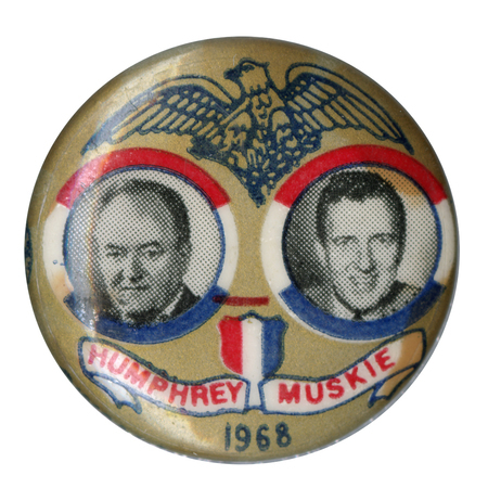 Round, gold button with portraits of Hubert Humphrey and Edmund Muskie surrounded by an illustration of an eagle and red, white, and blue shield and ribbon. Text reads "Humphrey Muskie 1968."