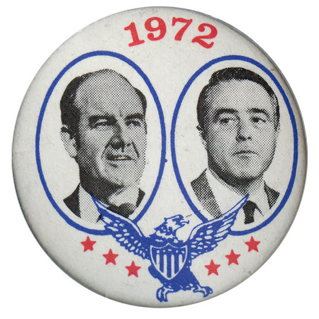 Round, white button with black and white portraits of George McGovern and Sargent Shriver in blue circles. Below the portraits are a blue eagle and red stars. Red text reads "1972"