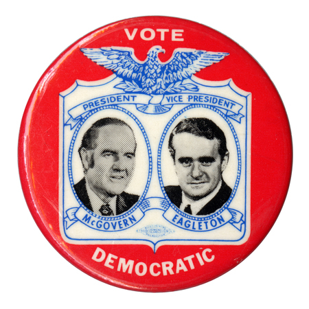Round, red button with black and white portraits of George McGovern and Sargent Shriver in an elaborate blue and white frame topped by an eagle. Text reads "VOTE DEMOCRATIC PRESIDENT McGOVERN VICE PRESIDENT EAGLETON"