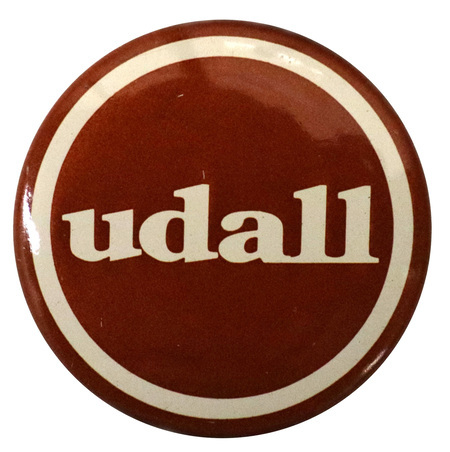Round, maroon and white button with white text reading "udall"