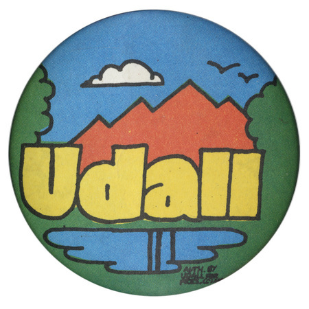 Round button with a green, red, blue, and white drawing of a mountain, trees, and lake. Yellow text reads "udall"