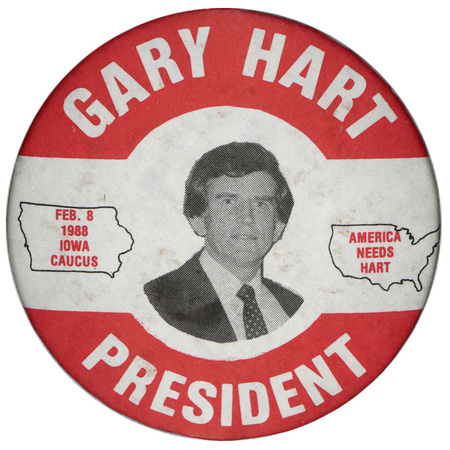 Round, white button with a red border and a black and white photograph of Gary Hart in the center. An outline of the state of Iowa contains red text reading "FEB. 8 1988 IOWA CAUCUS." An outline of the United States contains red text reading "AMERICA NEEDS HART." White text around the edge reads "GARY HART PRESIDENT"
