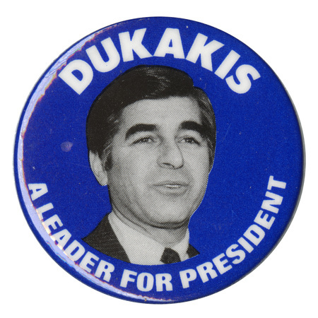 Round, white button with a black and white photograph of Michael Dukakis and white text reading "DUKAKIS A LEADER FOR PRESIDENT"