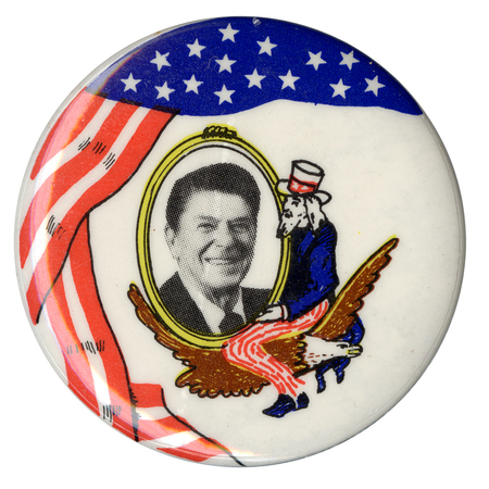 Round, white button with a red, white, and blue drawing of the US flag and a drawing of Uncle Sam sitting on an eagle and cradling an oval that contains a black and white photograph of Ronald Reagan.