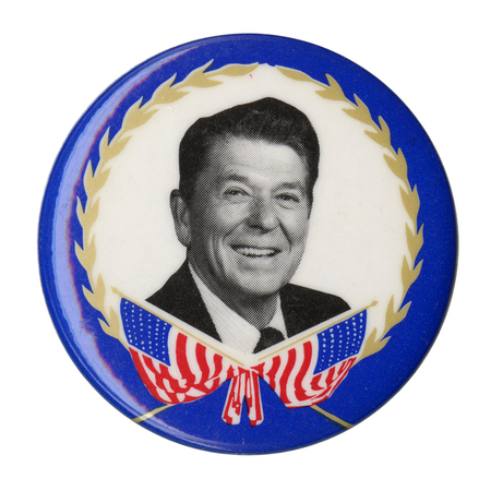 Round button with a blue border. Within a white circle in the center is a black and white photograph of Ronald Reagan above a drawing of two crossed US flags.