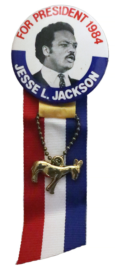 Round, white button with a black and white photograph of Jesse Jackson. Red and white text reads "FOR PRESIDENT 1984 JESSE L. JACKSON." Attached to the bottom of the button is a red, white, and blue ribbon and a gold donkey charm.