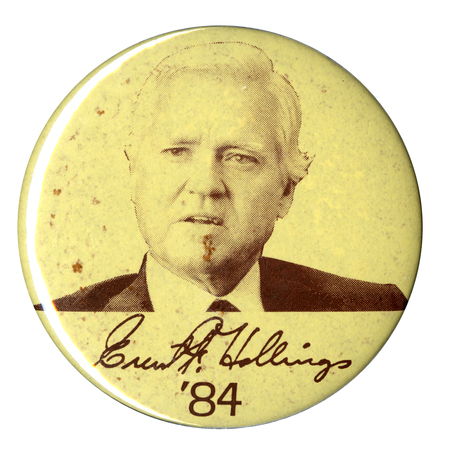 Round, off white button with a black and white photograph of Fritz Hollings. Under the photograph is Hollings's signature "Ernest F. Hollings" and "'84" in black. 