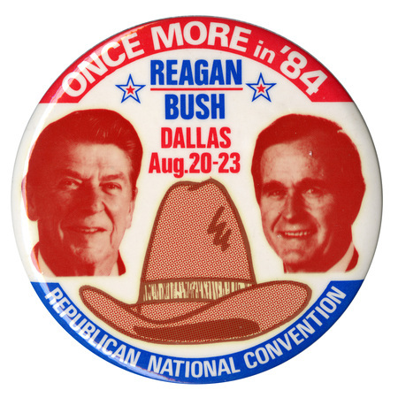 Round, white button with red and blue border. Red and white photographs of Ronald Reagan and George H. W. Bush appear on either side of a large drawing of a brown hat. White, red, and blue text reads "ONCE MORE in '84 REAGAN/BUSH DALLAS Aug. 20-23 REPUBLICAN NATIONAL CONVENTION"