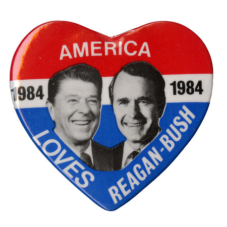 Heart-shaped button with bands of red, white, and blue and black and white photographs of Ronald Reagan and George H. W. Bush. White and black text reads "AMERICA 1984 LOVES REAGAN-BUSH"