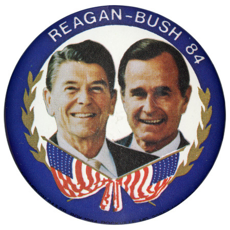 Round button with a blue border and color photographs of Ronald Reagan and George H. W. Bush surrounded by olive branches and two crossed US flags. White text reads "REAGAN-BUSH 84"