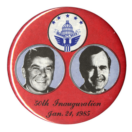 Round, red button with blue and white circles containing black and white photographs of Ronald Reagan and George H. W. Bush and a drawing of the US Capitol surrounded by an eagle bearing a shield, stars, and a ribbon with "REAGAN BUSH" on it. Black text below the photographs reads "50th Inauguration Jan. 21, 1985"