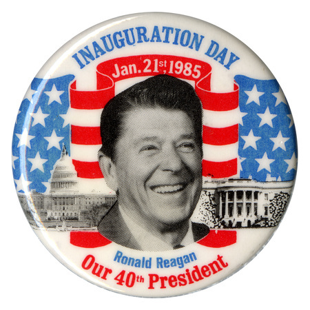 Round, white button with a black and white photograph of Ronald Reagan against black and white photographs of the US Capitol, the White House, and a drawing of the US flag in red, white, and blue. Blue, white, and red text reads "INAUGURATION DAY Jan. 21st, 1985, Ronald Reagan, Our 40th President"