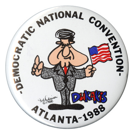 Round, white button with a caricature drawing of Michael Dukakis holding a US flag. Black text reads "DEMOCRATIC NATIONAL CONVENTION ATLANTA-1988"