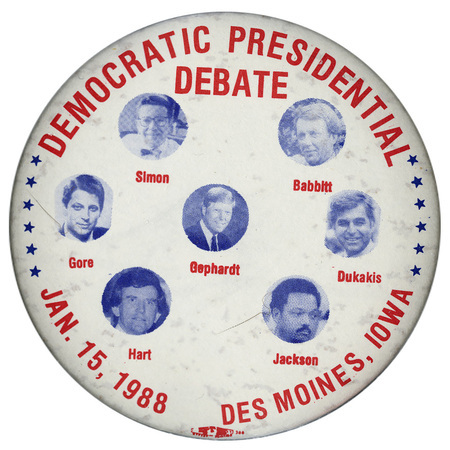 White button with black and white photographs of Paul Simon, Bruce F. Babbitt, Al Gore, Richard A. Gephardt, Michael Dukakis, Gary Hart, and Jesse Jackson, each with his name underneath his photograph. Red text reads "DEMOCRATIC PRESIDENTIAL DEBATE JAN. 15, 1988 DES MOINES, IOWA"