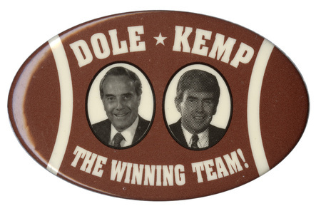 Oval button decorated in brown and white to look like an American football. In the center are black and white photographs of Bob Dole and Jack Kemp. White text reads "DOLE KEMP THE WINNING TEAM!"