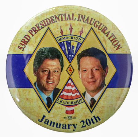 Round button with a splotchy yellow background and a band of blue across the middle. Color photographs of Bill Clinton and Al Gore appear inside interlocked diamond shapes in the center. Between the photographs is an illustration of a US flag. Red and black text reads "53RD PRESIDENTIAL INAUGURATION DEMOCRATIC LEADERSHIP January 20th"