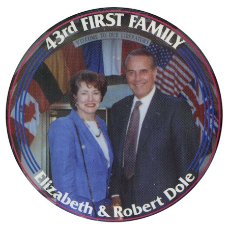 Round button with a border of red and blue stripes and a color photograph of Elizabeth and Bob Dole in front of a series of national flags and a sign reading "WELCOME TO OUR LIBERATORS". White text on the button reads "43rd FIRST FAMILY Elizabeth & Robert Dole"
